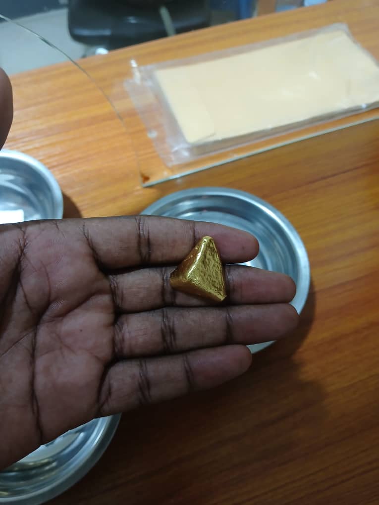 Triangular gold doré button resting on fingers above a stainless assay dish